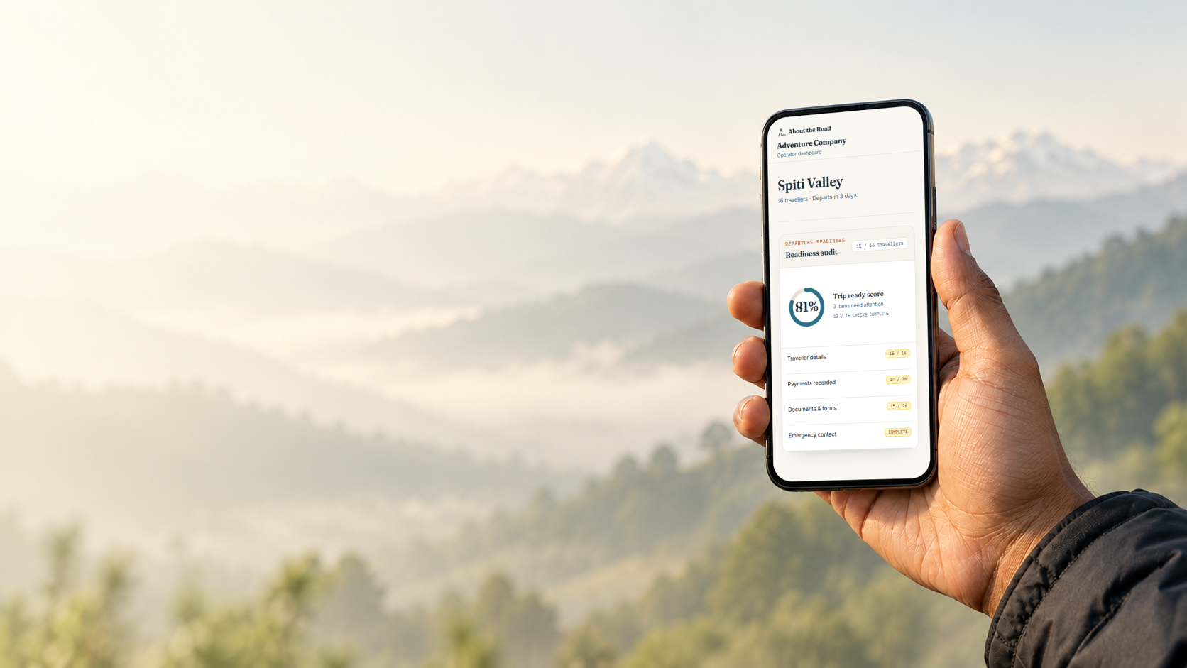 A trip operator's hand holding up a phone showing the About the Road trip-ready dashboard, misty forested hills in the background